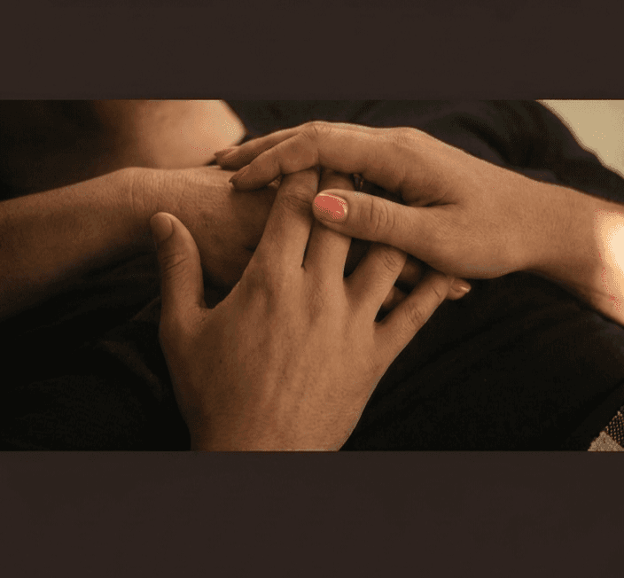 Two people's hands clasped together over a chest, one hand featuring bright pink painted fingernails.